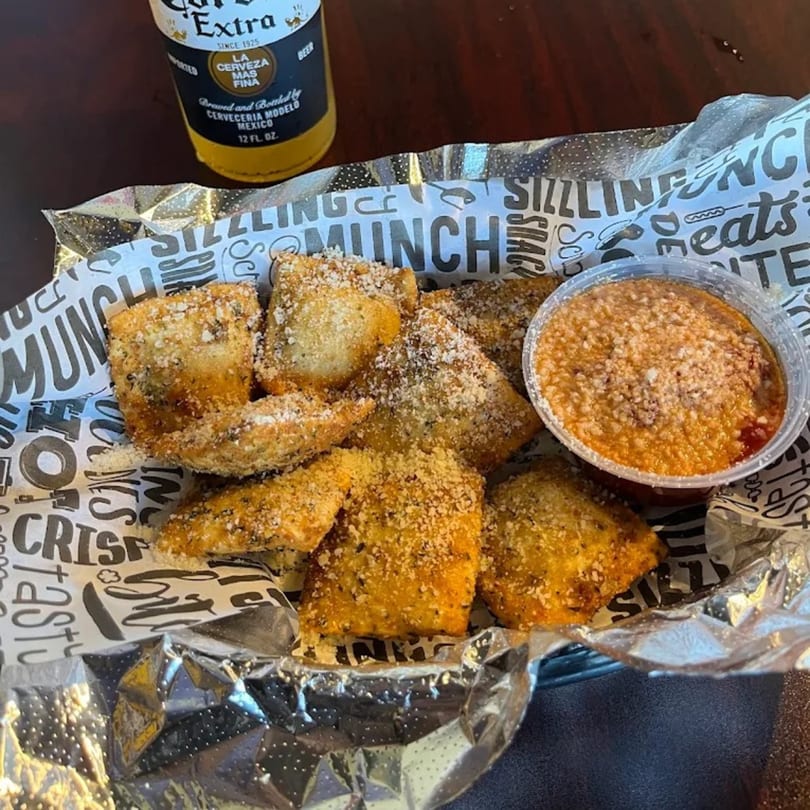 Toasted ravioli with marinara sauce and a Corona Extra beer on a dark table.
