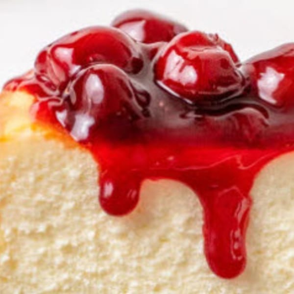 Close-up of a slice of cheesecake with bright red cherry topping dripping down the side.